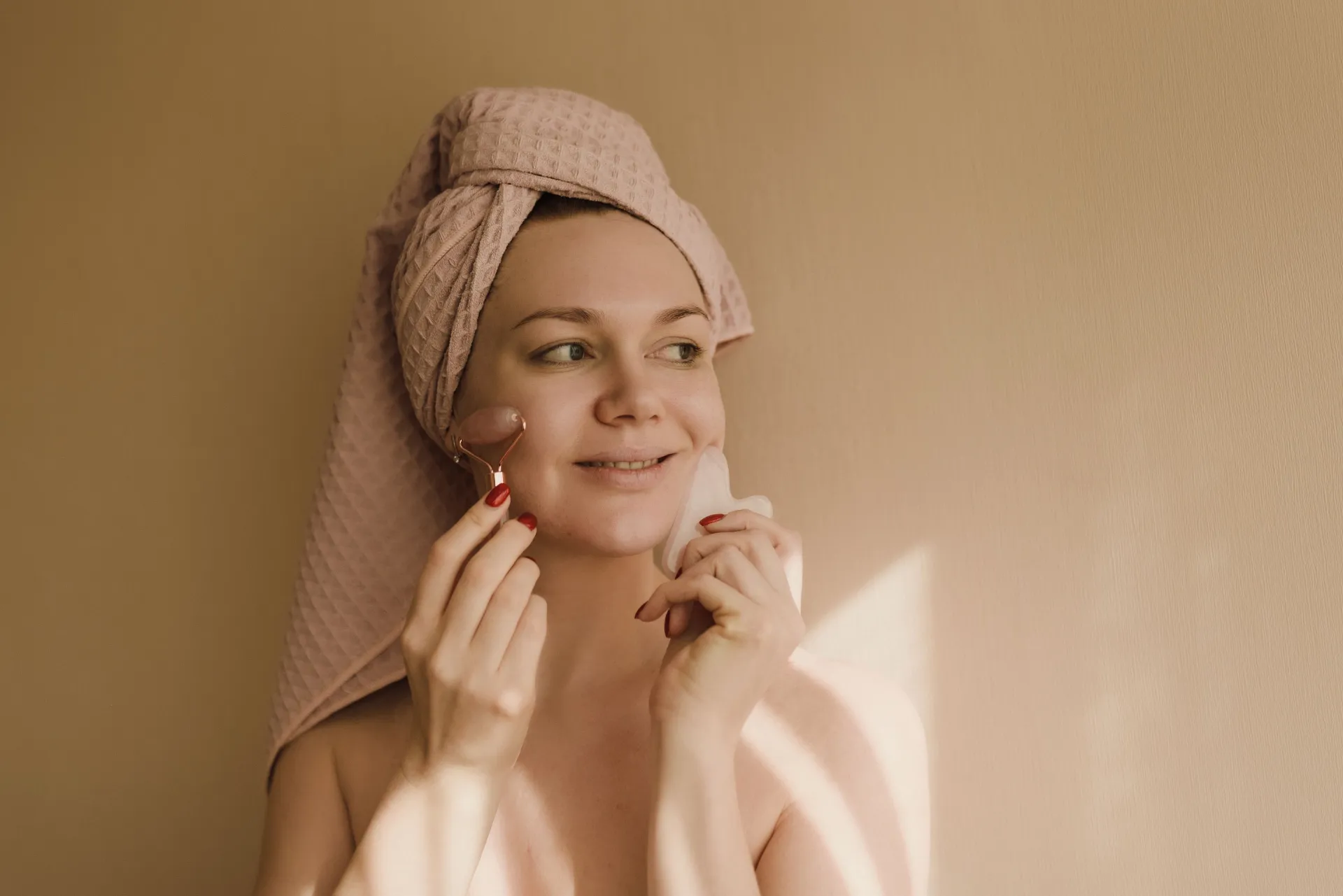 Woman in her 30s smiling during a calm gua sha and facial roller skincare routine in soft, warm natural light.