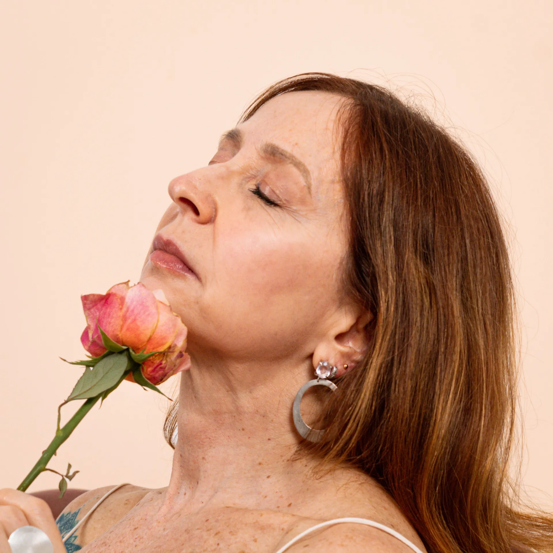 Serene mature woman with radiant, naturally textured skin holding a pink rose — calm, confident, refreshed not overdone.