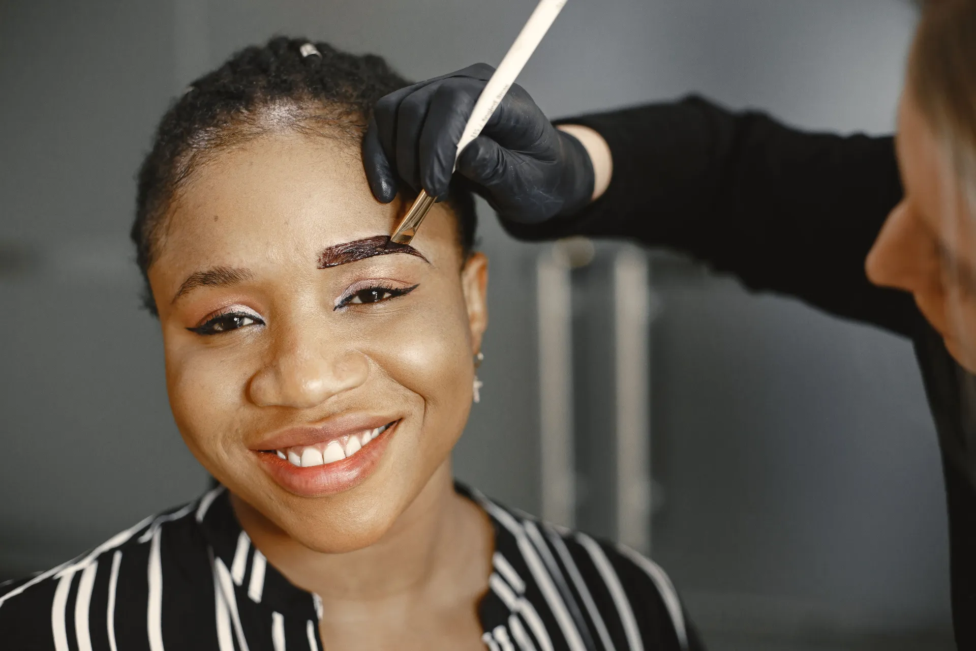 Beautician applying a custom eyebrow tint with an angled brush as a smiling client relaxes during a brow wax and tint visit.