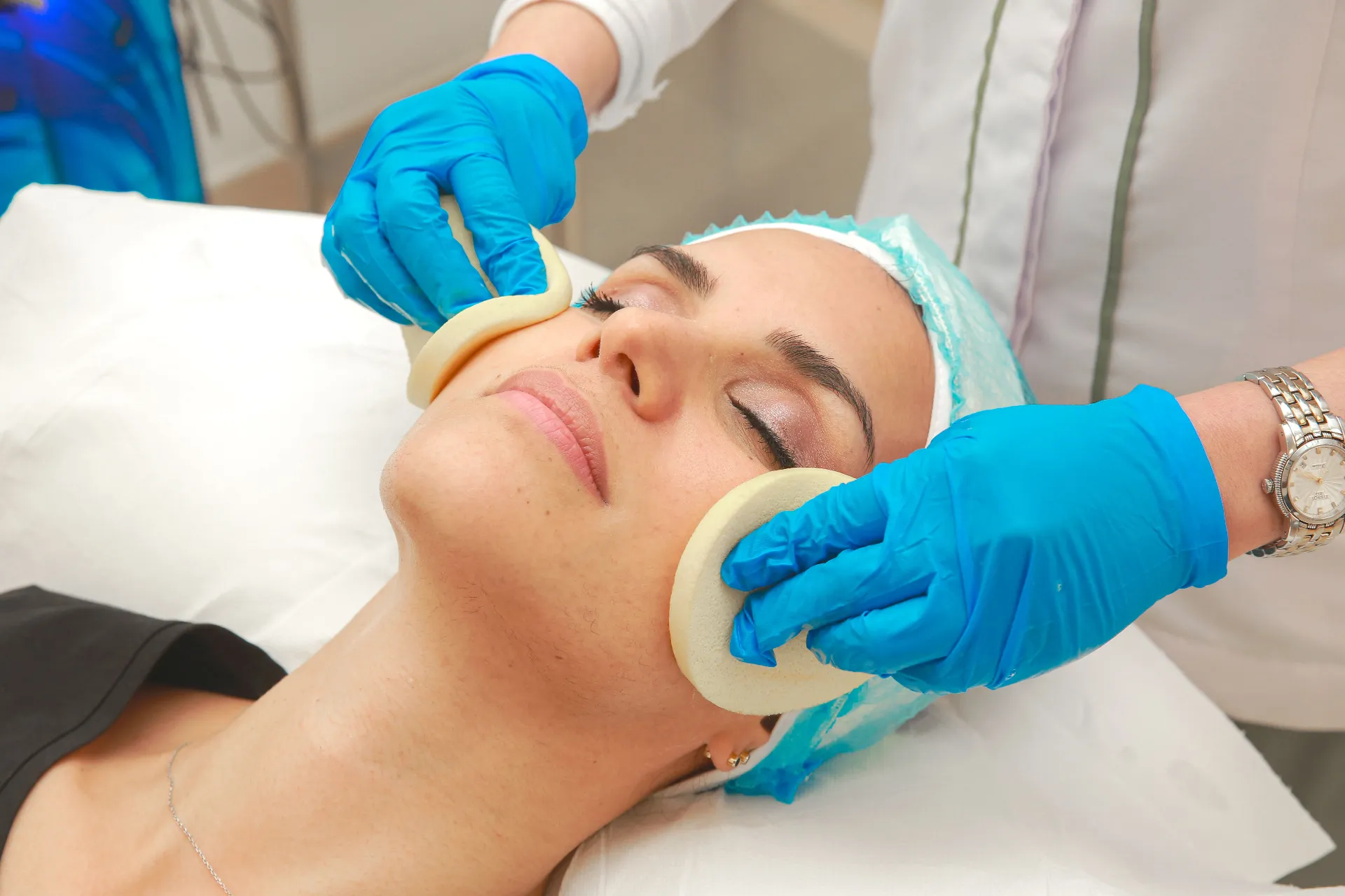 Esthetician exfoliating a client's skin with cleansing sponges during a professional hydrating facial in a calm treatment room.