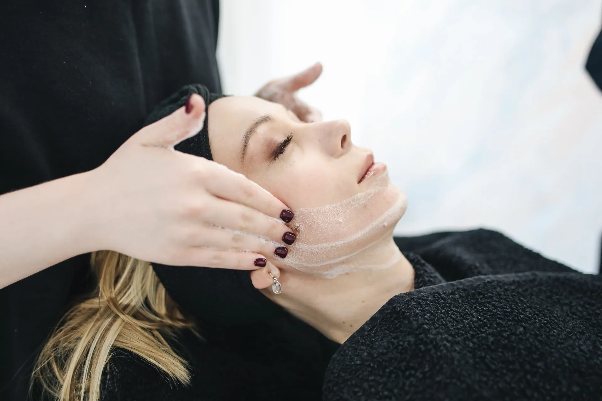 Esthetician smoothing an exfoliating treatment along a woman's jawline as she relaxes during a calming spa facial.